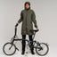 woman wearing a green tench raincoat holding a black Brompton folding bicycle 