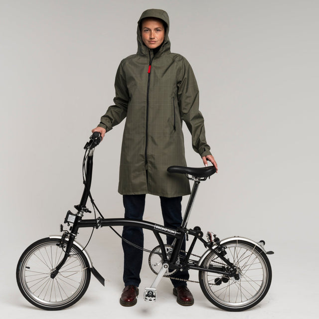 woman wearing a green tench raincoat holding a black Brompton folding bicycle 